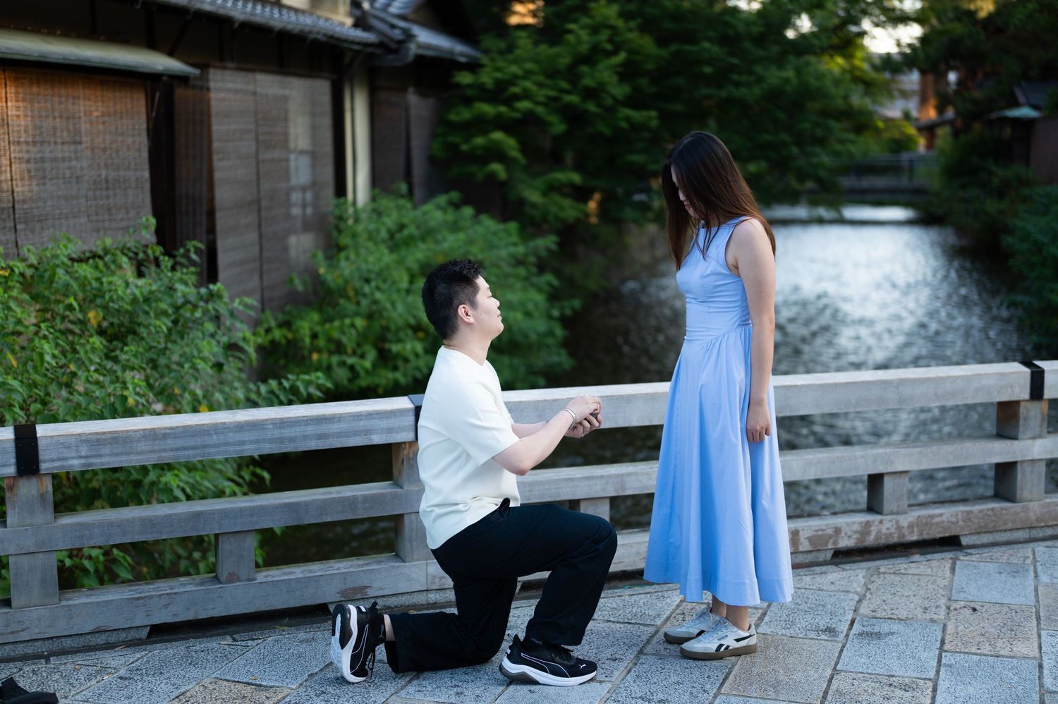 Man in white shirt kneels on one knee proposing to a woman in a blue dress on a stone bridge beside a calm river.