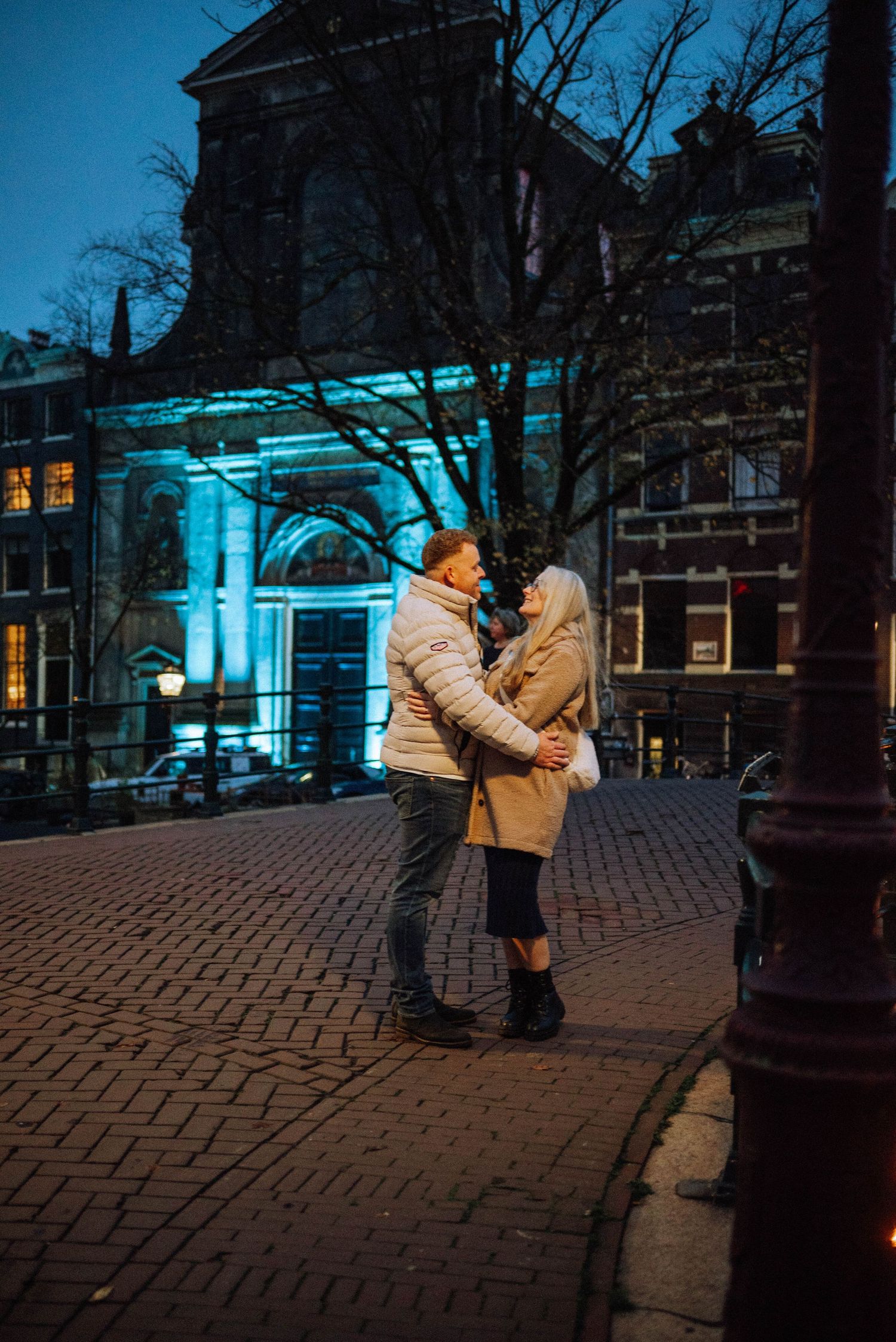 Couple in winter jackets embracing on a brick street at dusk in front of a historic building glowing with light. Reguliersgracht Amsterdam