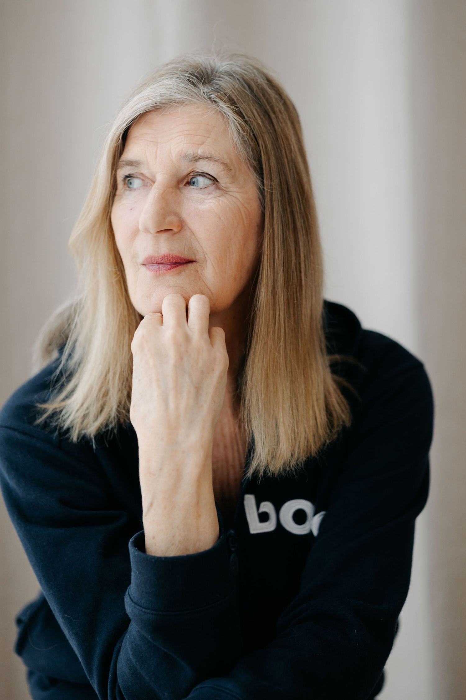 Thoughtful middle-aged blonde woman in black hoodie resting chin on hand, gazing upward against neutral background.
