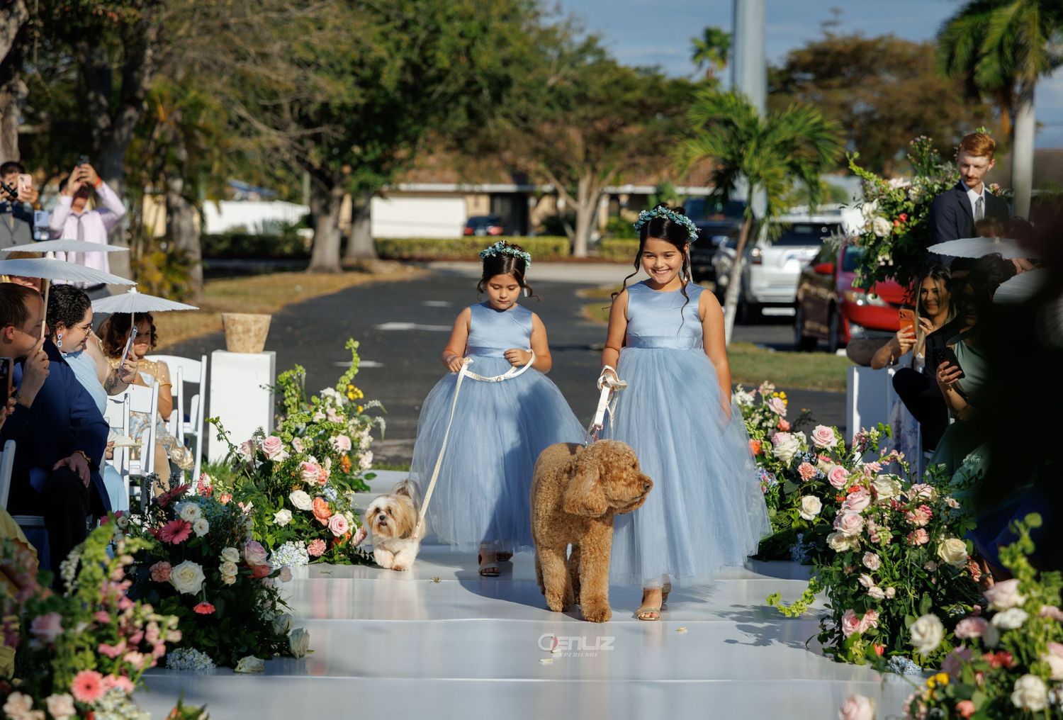 Two flower girls in blue dresses walk a dog down a floral wedding aisle outdoors.