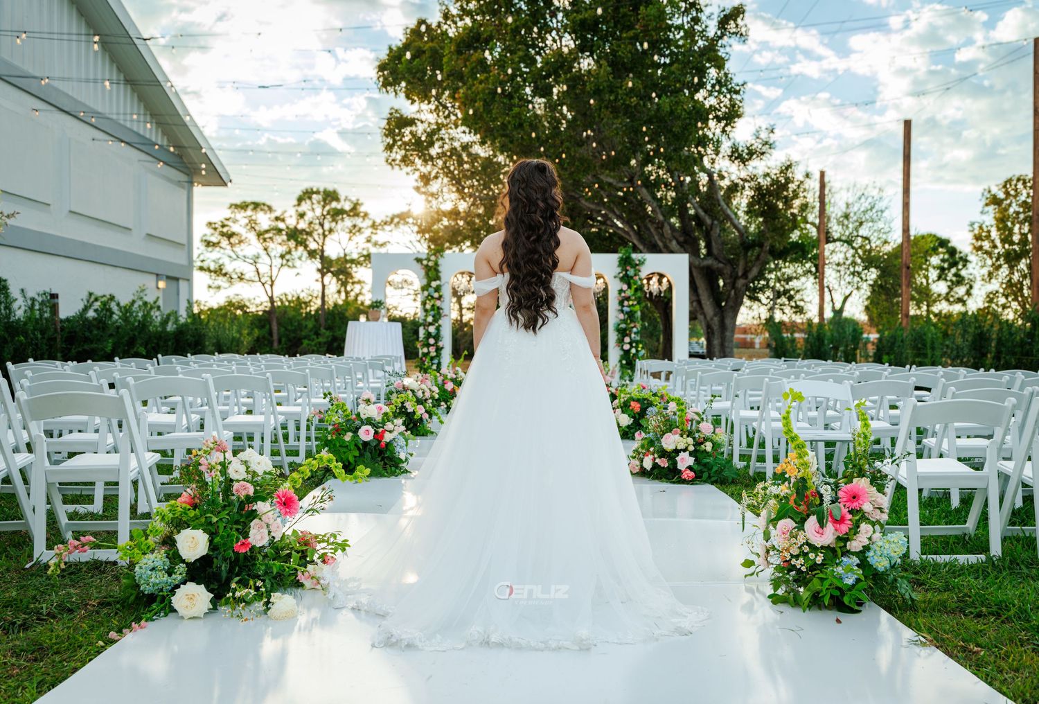 Bride in white gown stands at floral-lined aisle with white chairs and arch at outdoor wedding ceremony.