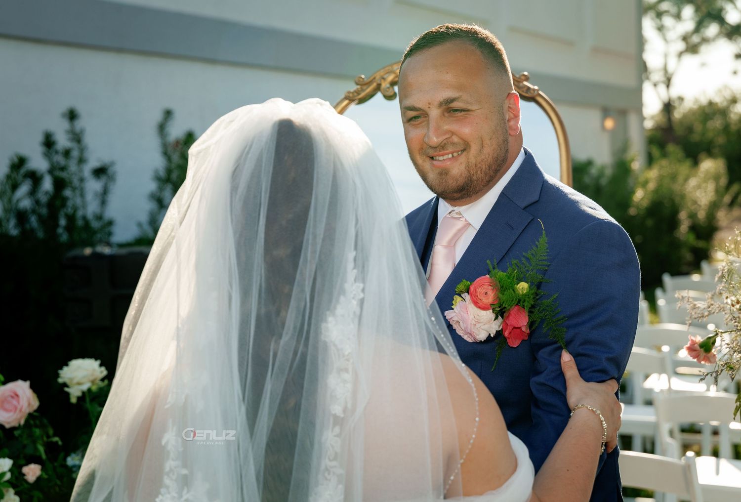 Groom in navy suit smiling at bride in white lace dress and veil during outdoor wedding ceremony.