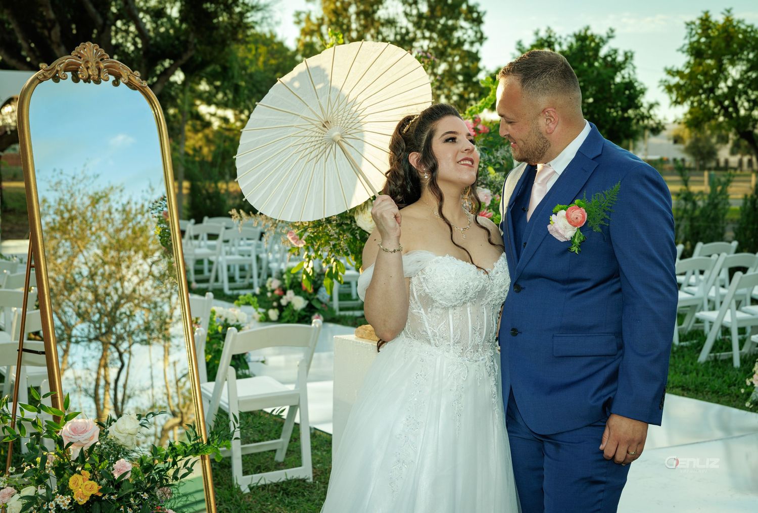 Bride and groom pose outdoors at wedding ceremony, bride holding white parasol beside ornate gold mirror.