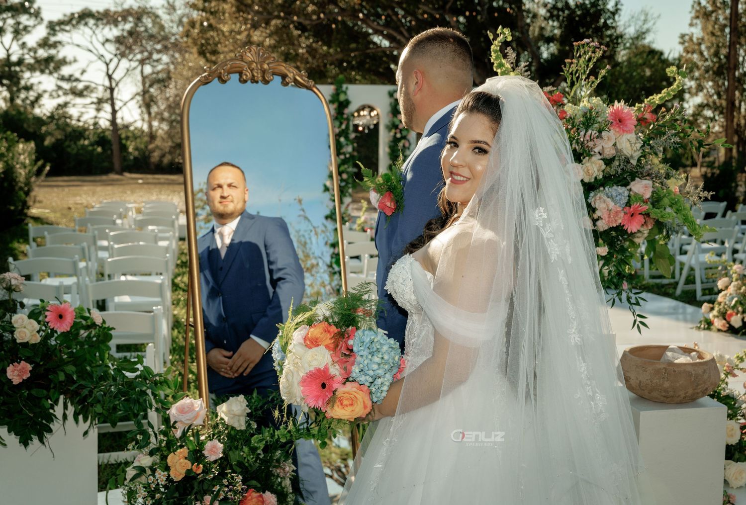 Bride in white gown and veil holds colorful bouquet beside gold mirror reflecting groom in blue suit at outdoor wedding.