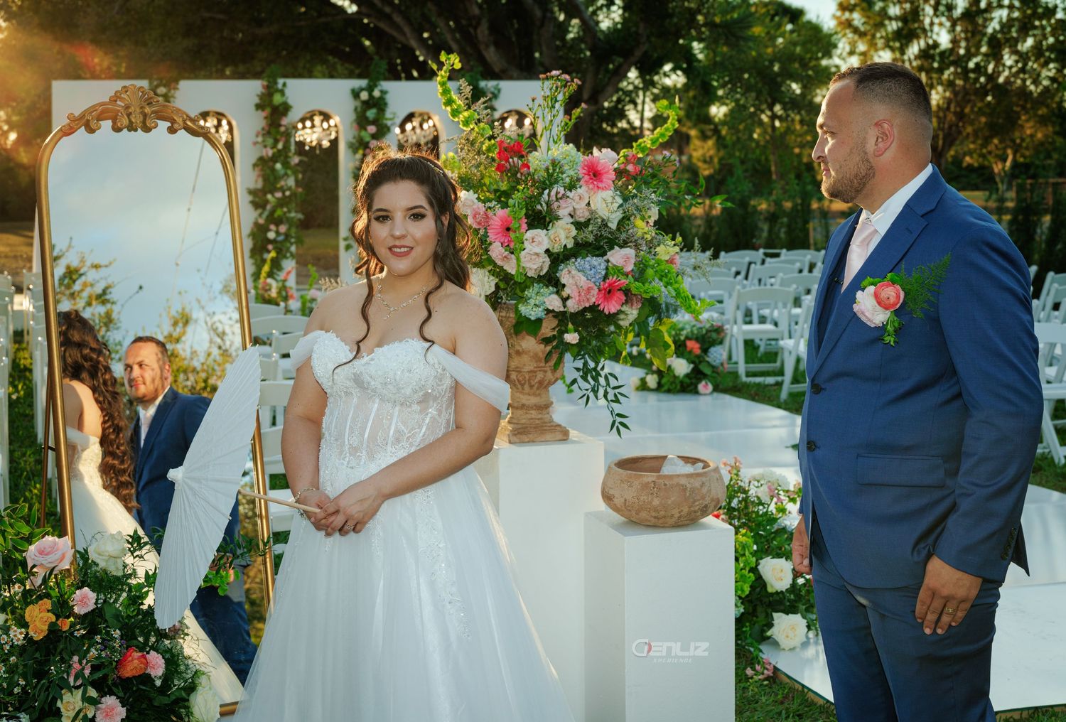 Bride in white gown and groom in blue suit stand at outdoor wedding altar with colorful floral arrangements.