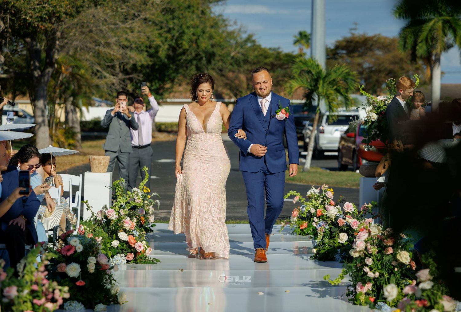 A couple walks down an outdoor wedding aisle lined with floral arrangements, the woman in lace and man in a blue suit.