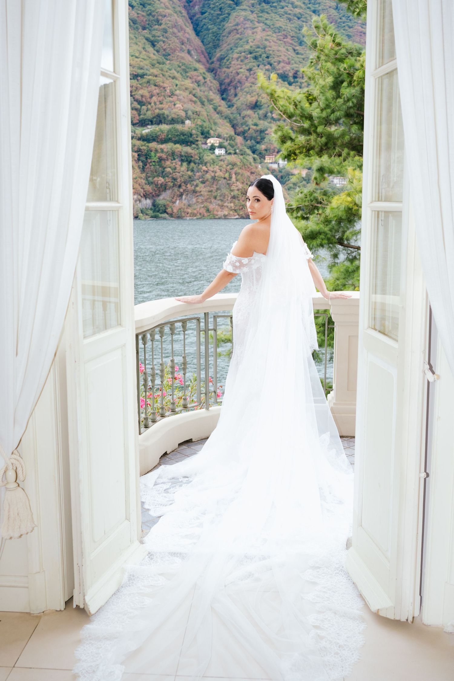 Bride in white off-shoulder gown and long veil stands on balcony overlooking scenic lake and mountains.