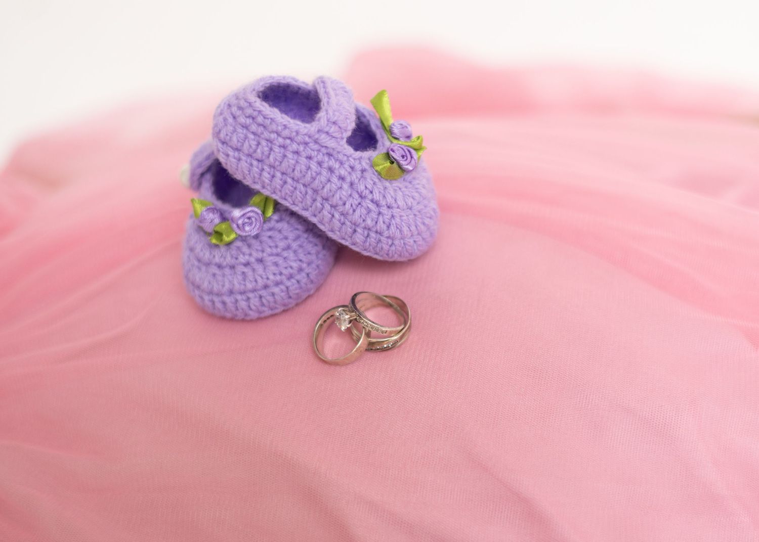 Purple crocheted baby booties with floral details beside wedding rings on pink tulle fabric.
