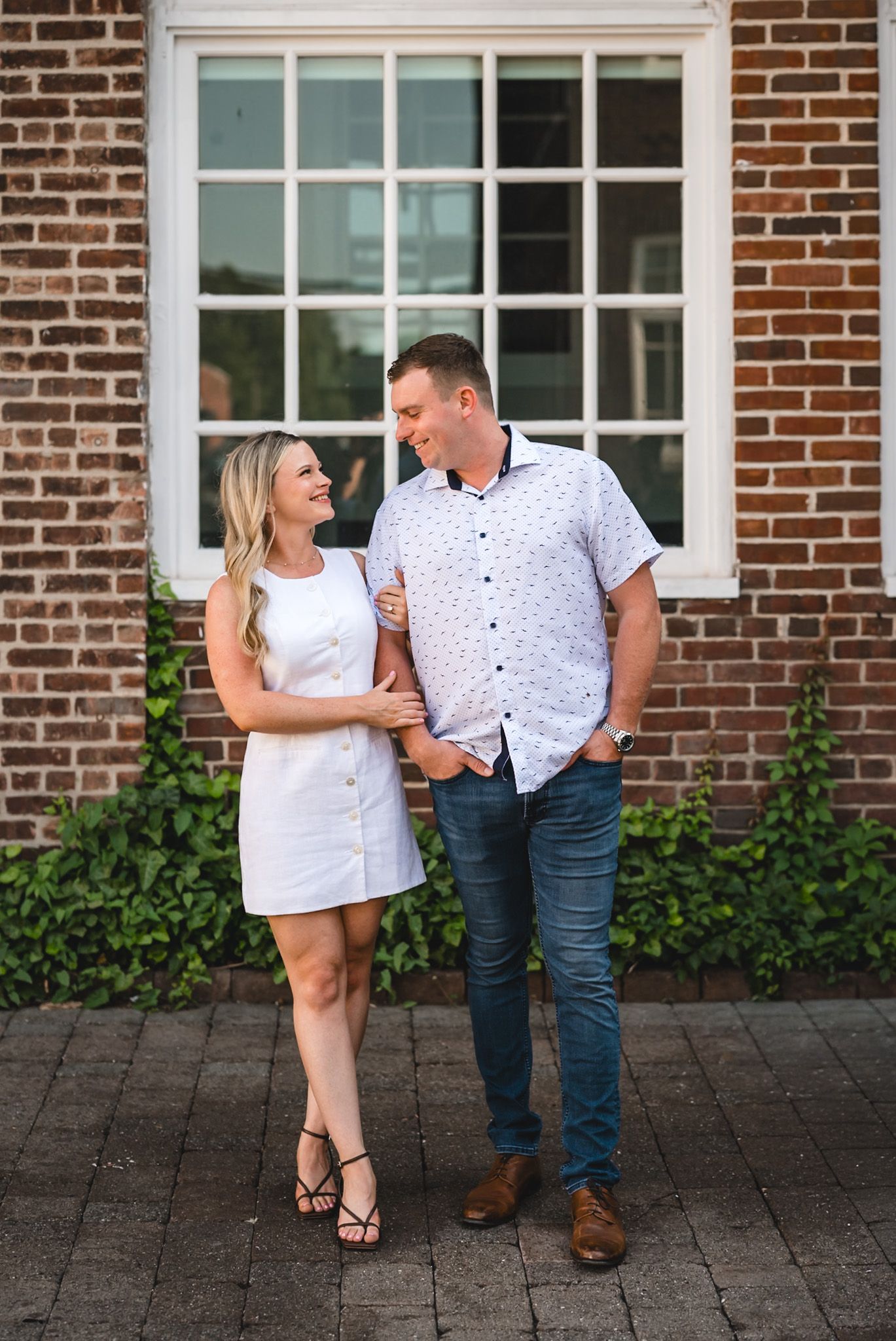 Couple smiling at each other in front of brick building, woman in white dress, man in blue shirt and jeans.