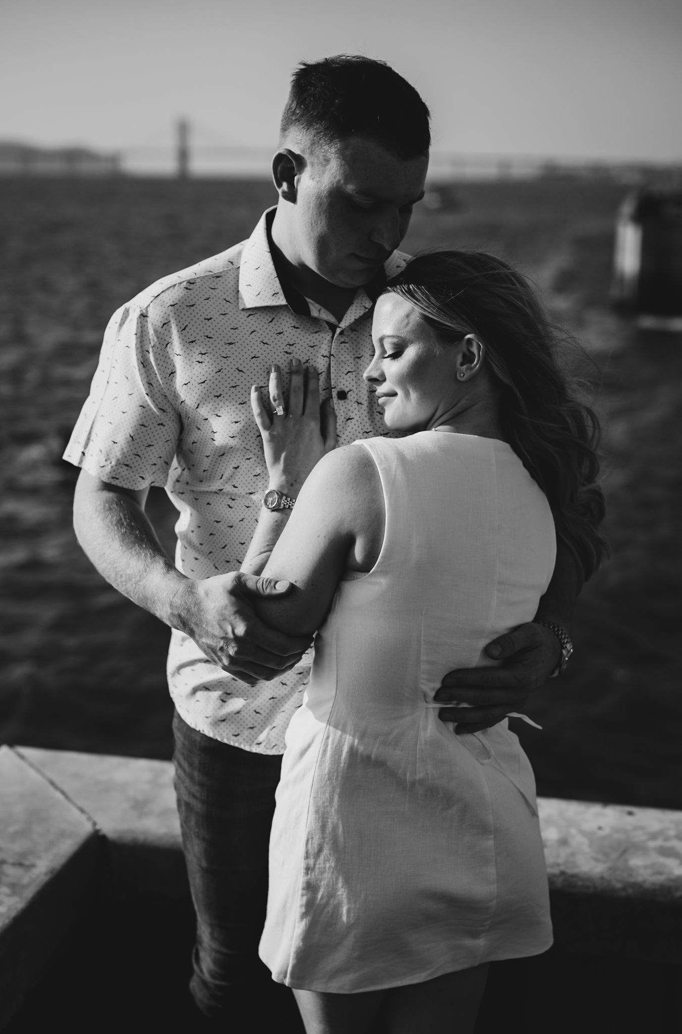 Black and white photo of a couple embracing outdoors near a waterfront, woman in white dress, man in patterned shirt.