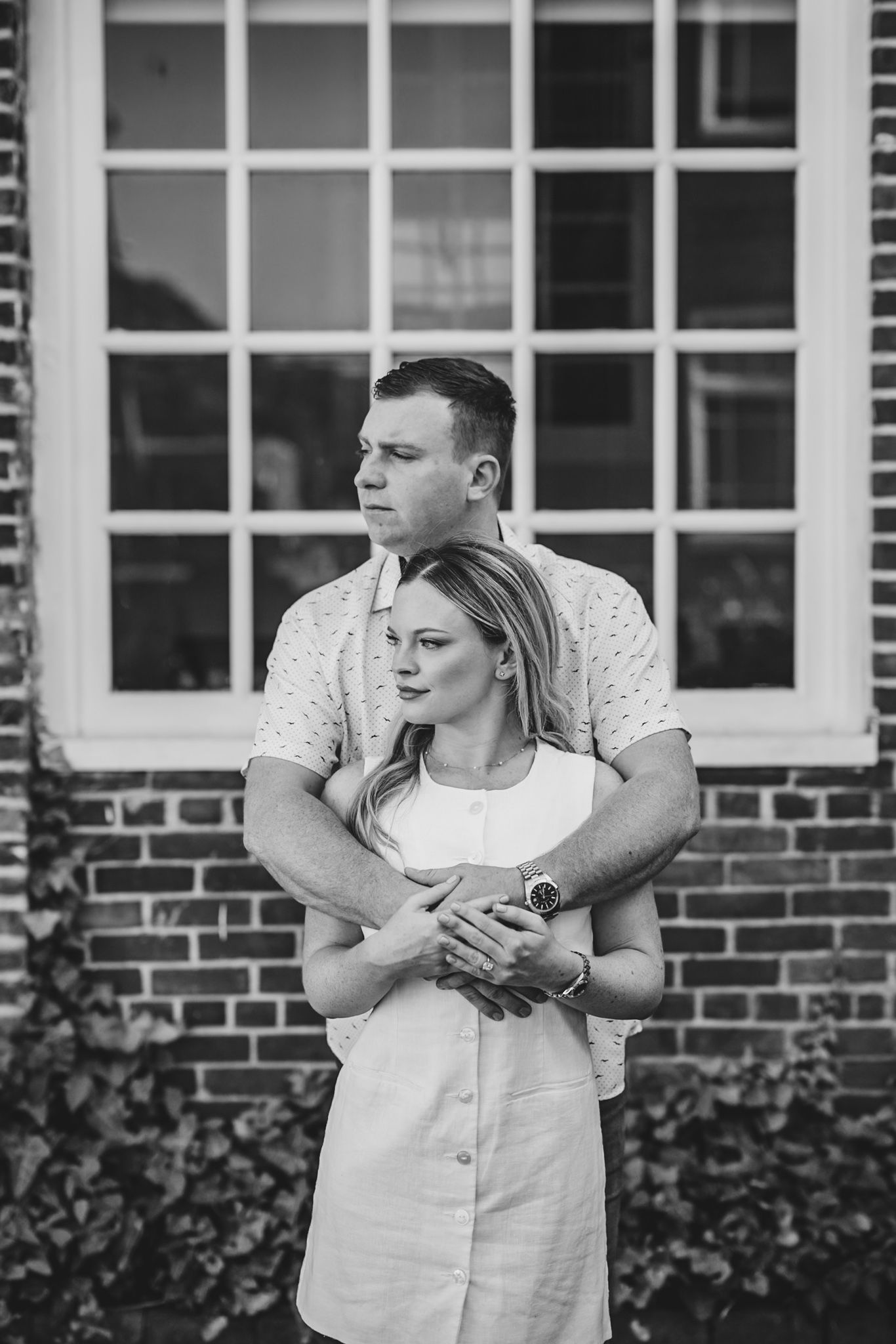 Black and white photo of a couple embracing in front of a brick wall and window.