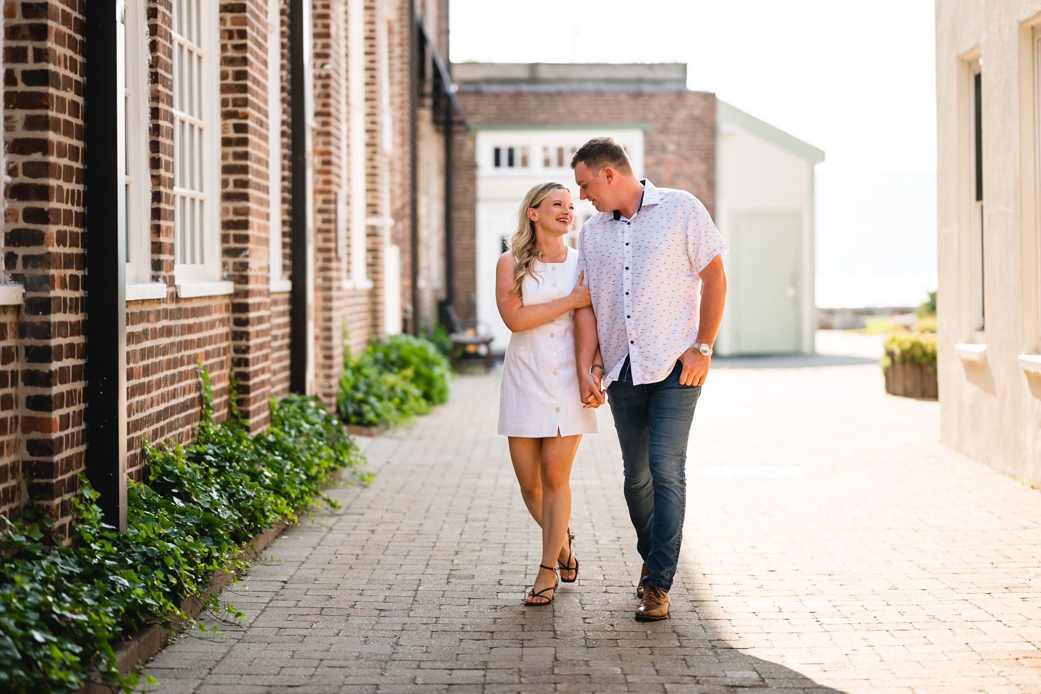Couple walking hand-in-hand along a brick pathway beside historic buildings, smiling at each other on a sunny day.