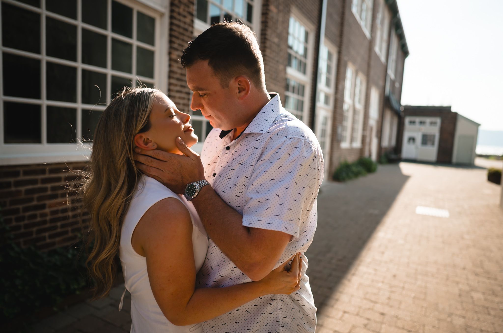 Couple sharing a romantic moment outdoors near a brick building, man gently holding womans face in golden sunlight.