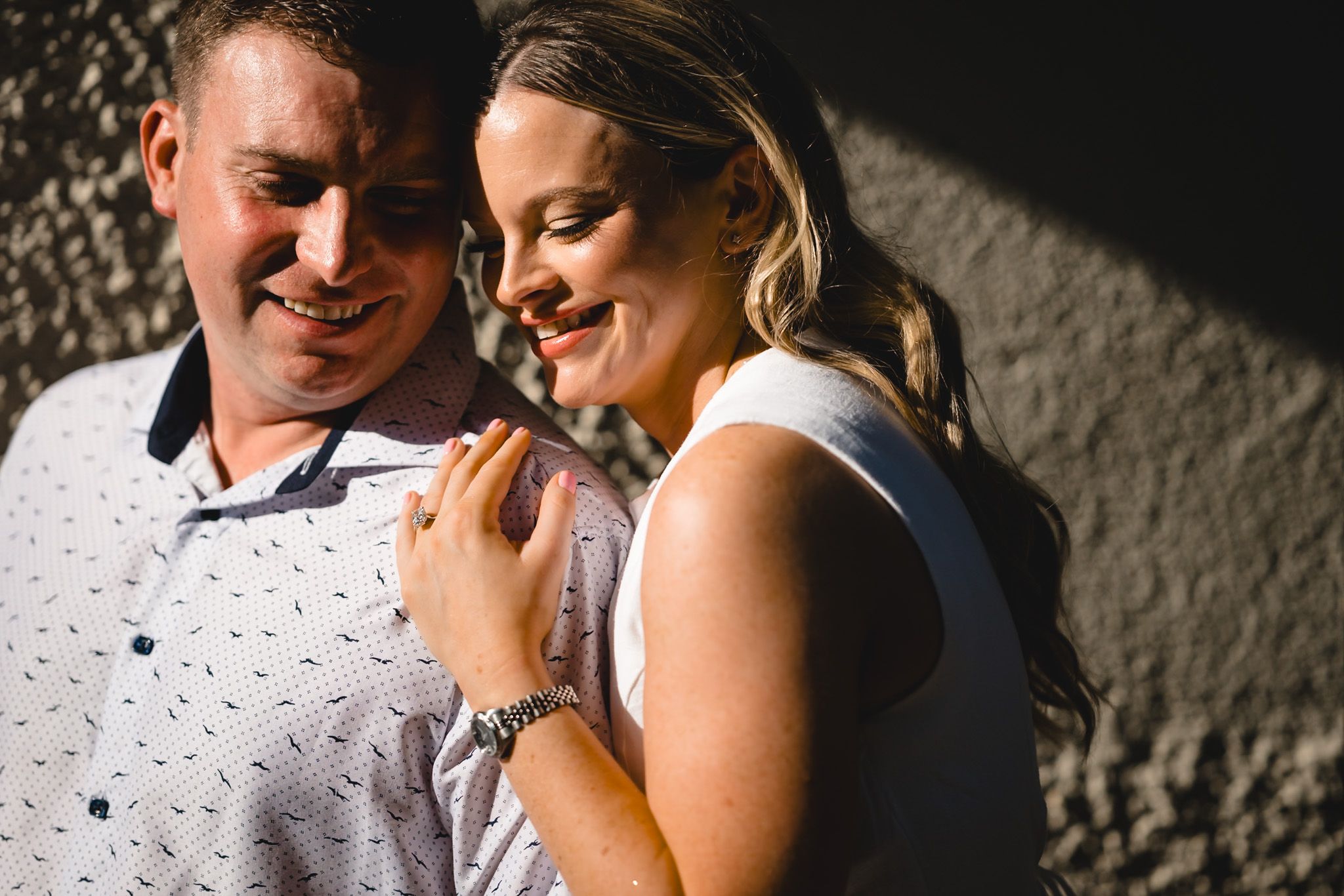 A couple shares a romantic moment, the woman smiling as she leans into the man against a textured wall.