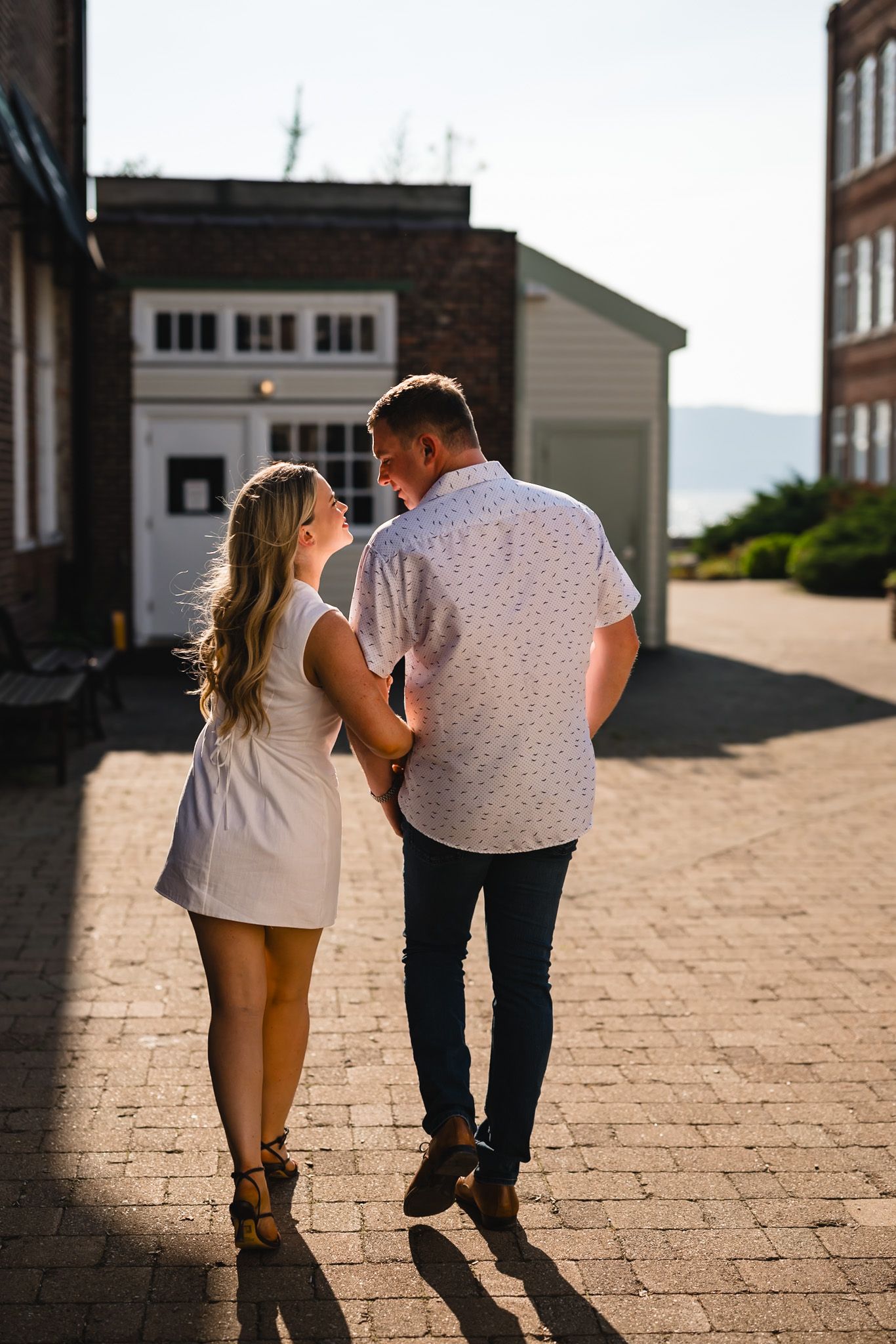 Couple walking hand in hand on cobblestone path, woman in white dress, man in patterned shirt, golden hour light.