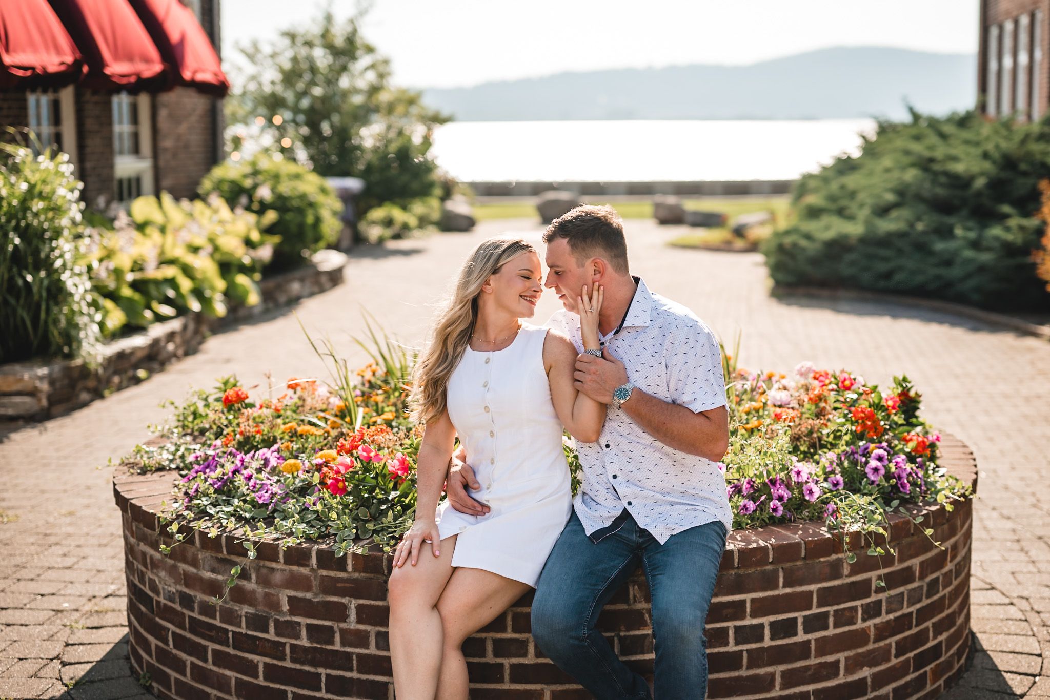 Couple sitting together on a brick planter surrounded by colorful flowers with a scenic waterfront view in the background.