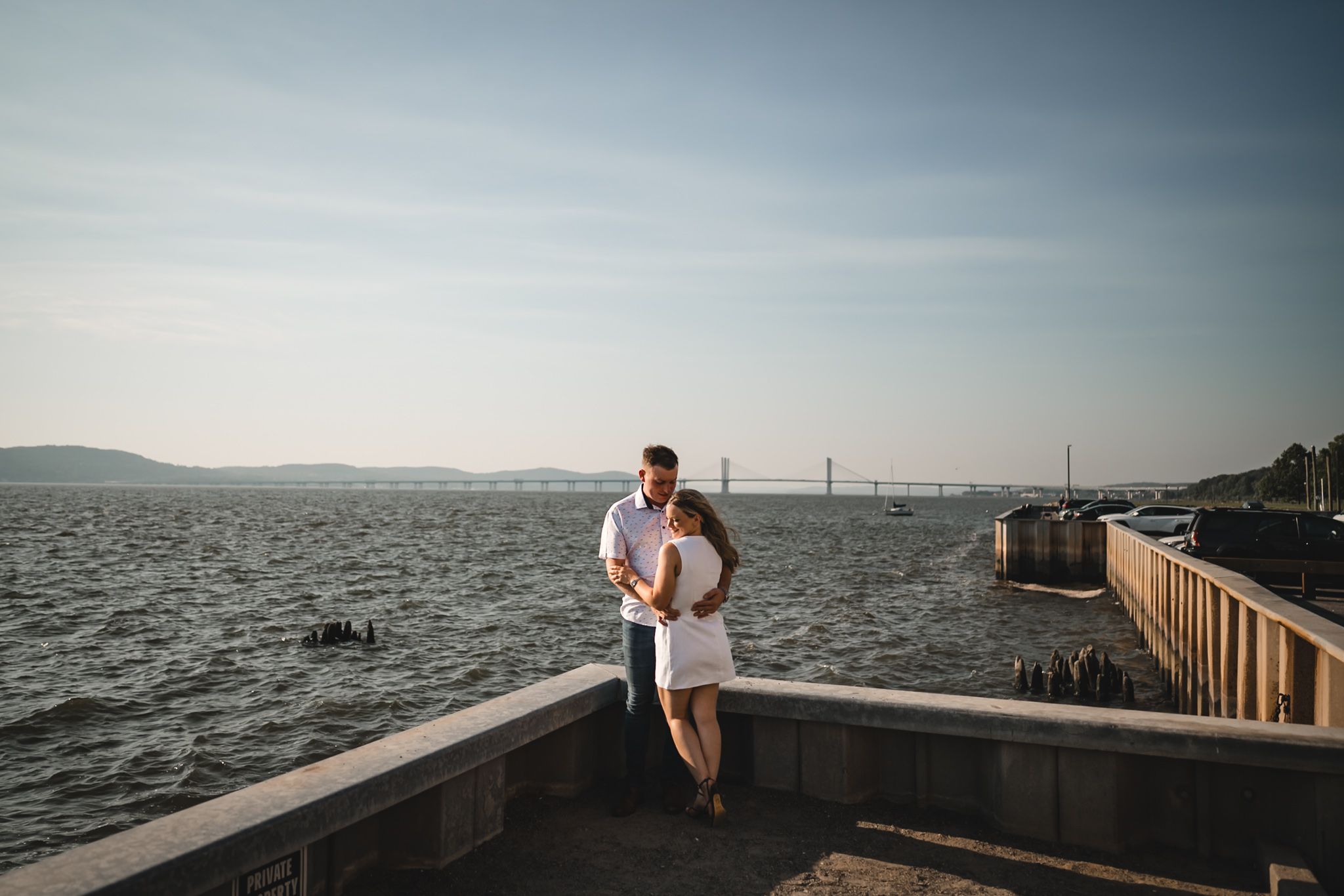 Couple embracing on a waterfront pier with a bay bridge and calm waters in the background on a sunny day.