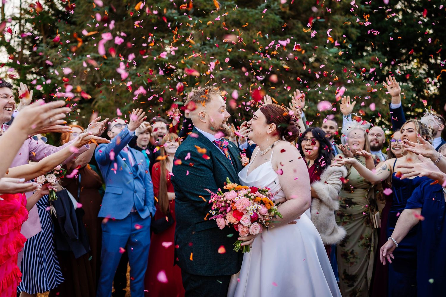Bride and groom embrace as wedding guests cheer and throw pink and red confetti petals outdoors at The River Barn by Lynette Matthews Photo
