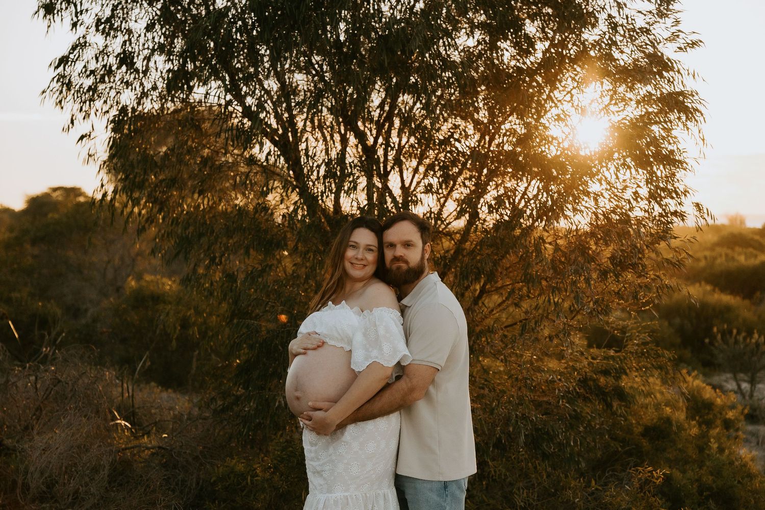 Pregnant woman in a white dress embraces her bearded partner outdoors at sunset, surrounded by trees and golden light.