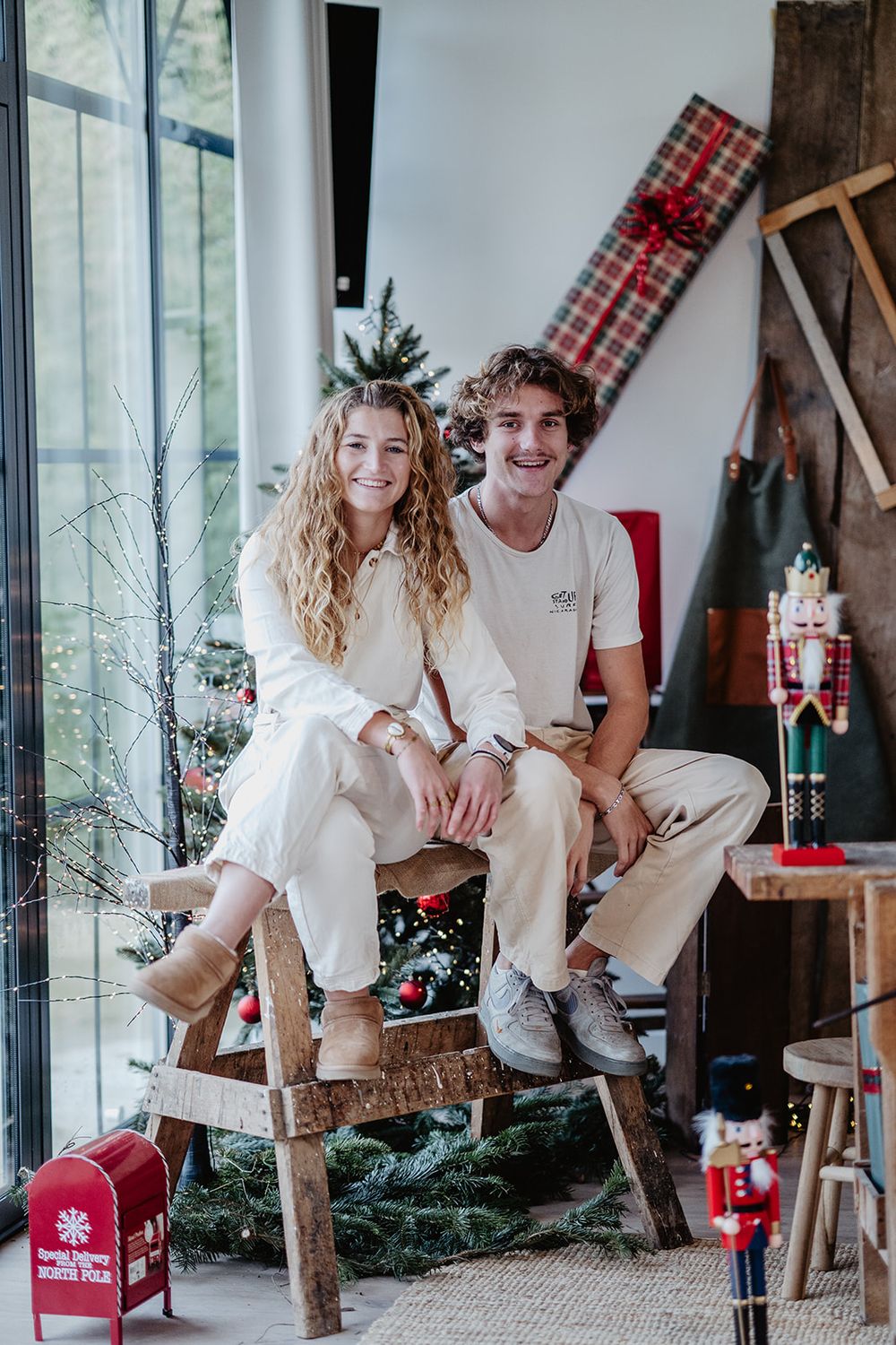 Young smiling couple in matching cream outfits sitting on wooden bench in festive Christmas decorated studio setting.