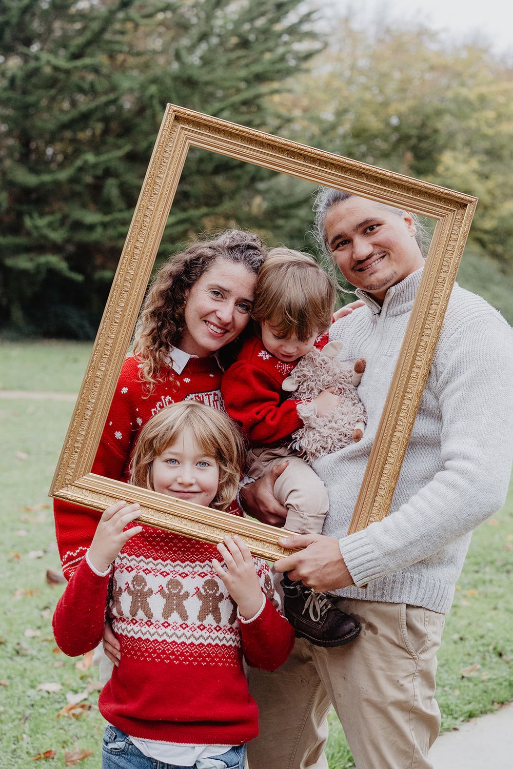 Family of four posing with a gold picture frame outdoors in Christmas sweaters.