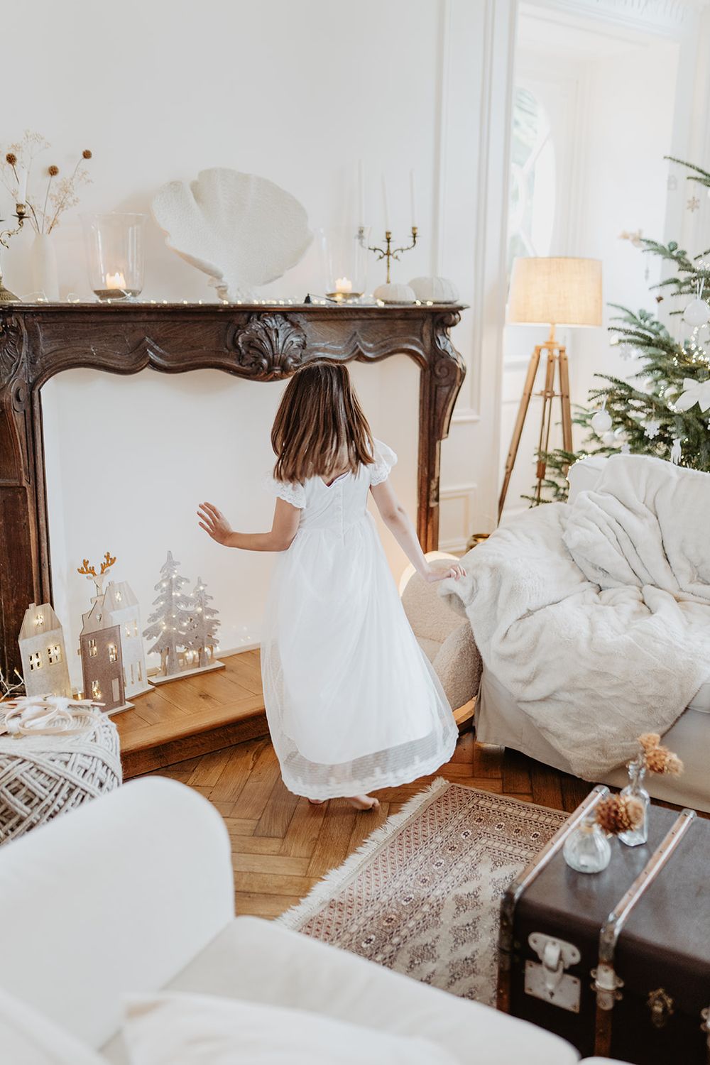 Young girl in white dress running toward ornate fireplace in elegant Christmas room.