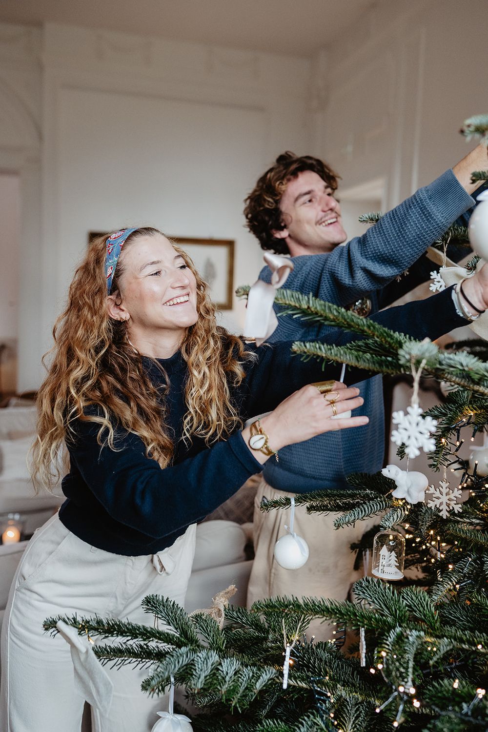 Smiling couple in navy sweaters happily decorating a Christmas tree with white ornaments in a bright living room.