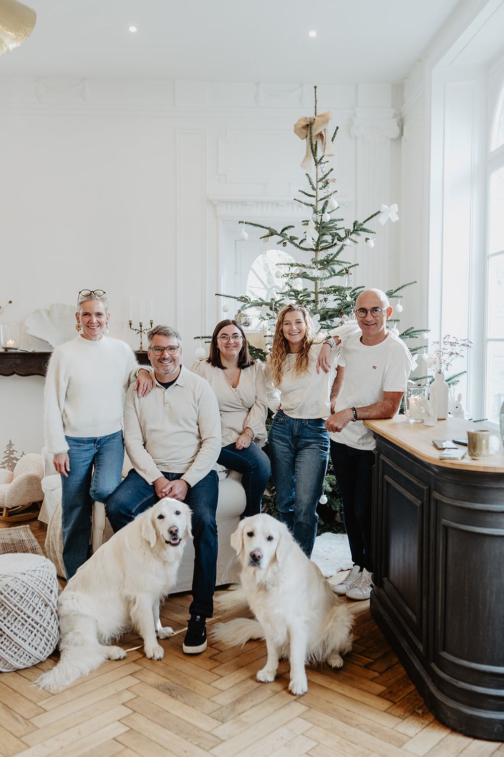Family of five with two golden retrievers posing in bright white room with minimalist Scandinavian Christmas tree decor.