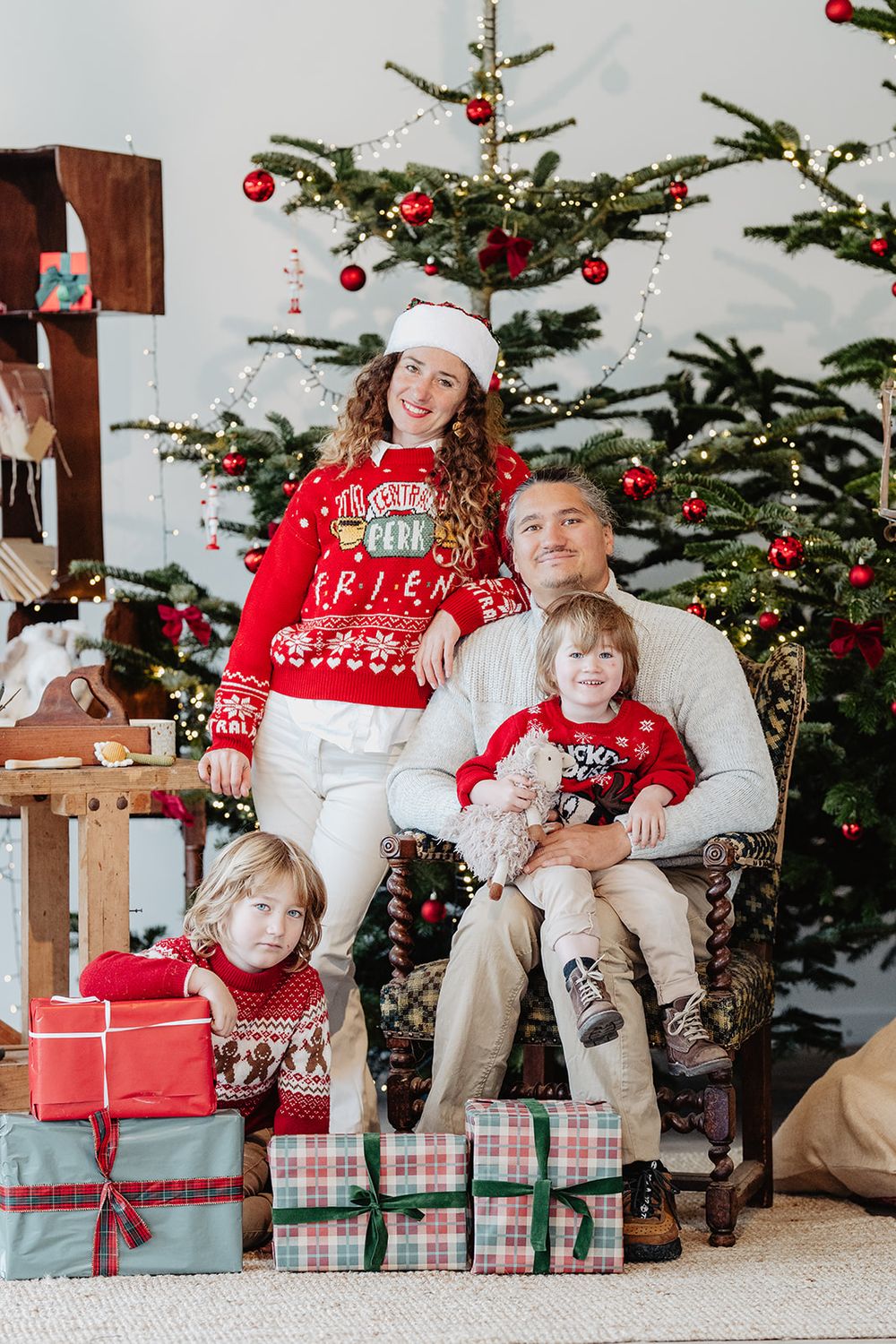 Happy family of four in red and white Christmas sweaters posing with gifts in front of decorated holiday trees.