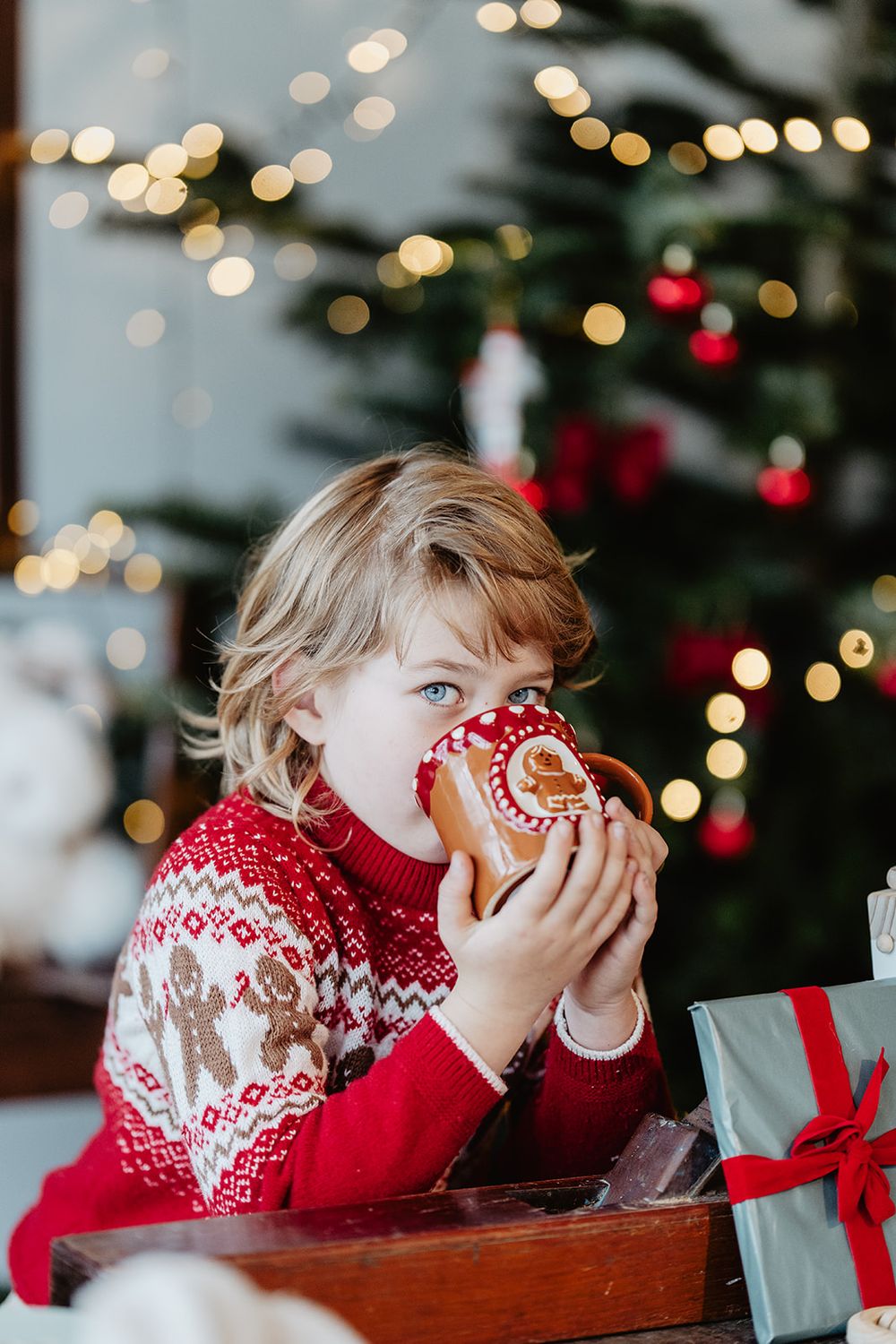 Blonde child in red Nordic sweater eating gingerbread cookie near Christmas tree with bokeh lights and wrapped gifts.