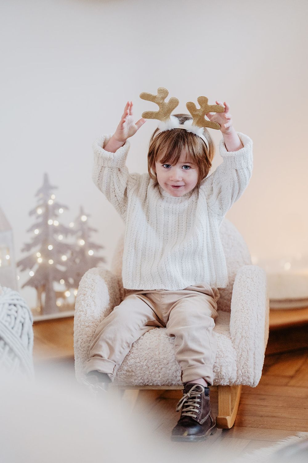 Cute child wearing reindeer antlers sits in a fluffy chair with Christmas decor.