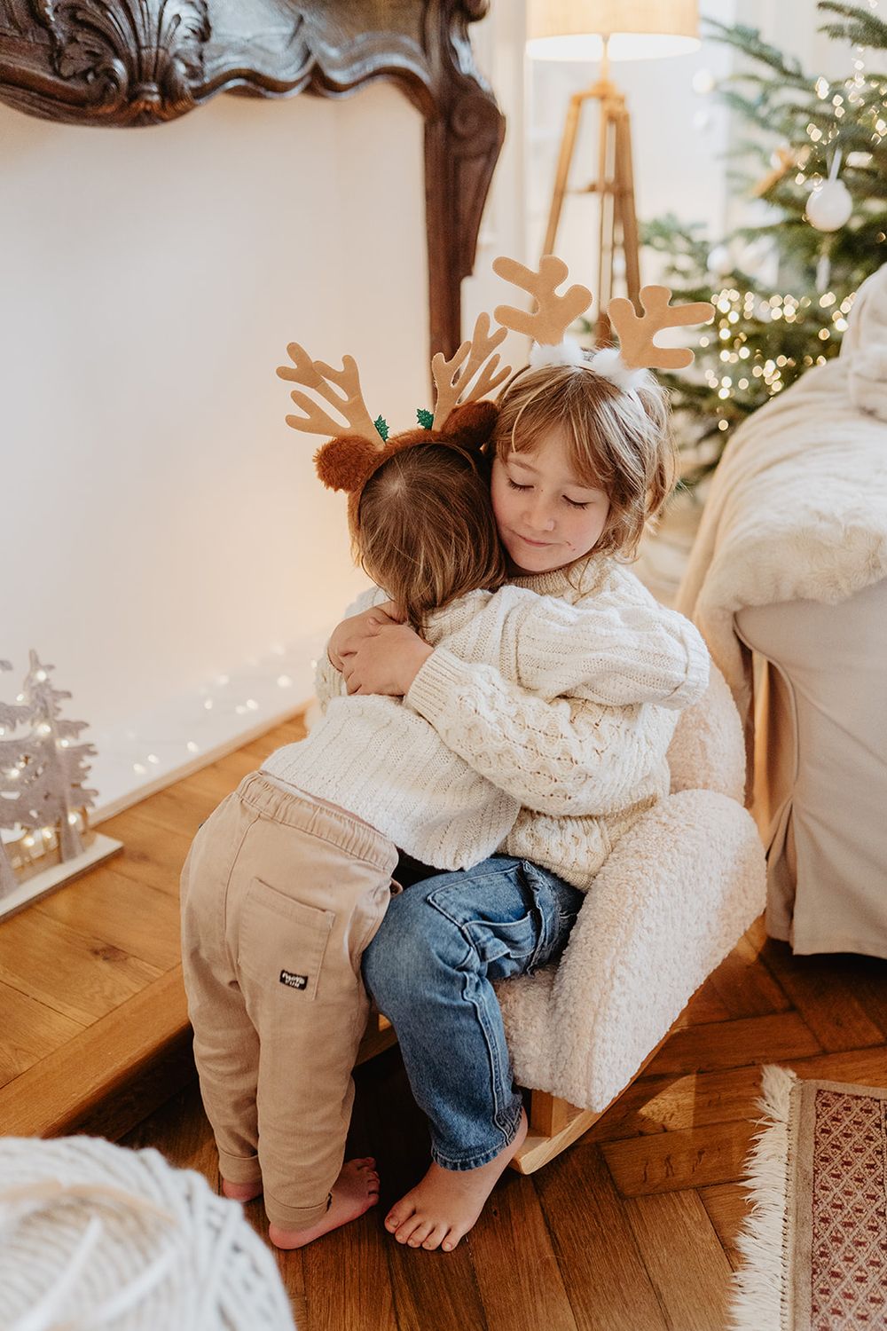 Two young children in cozy sweaters sharing a warm hug beside a glowing Christmas tree with reindeer antler headband.