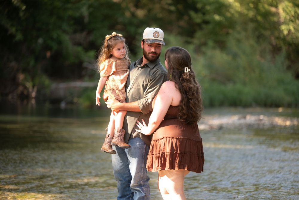 A young family stands in a shallow river, father holding toddler daughter while mother looks on, surrounded by lush greenery.