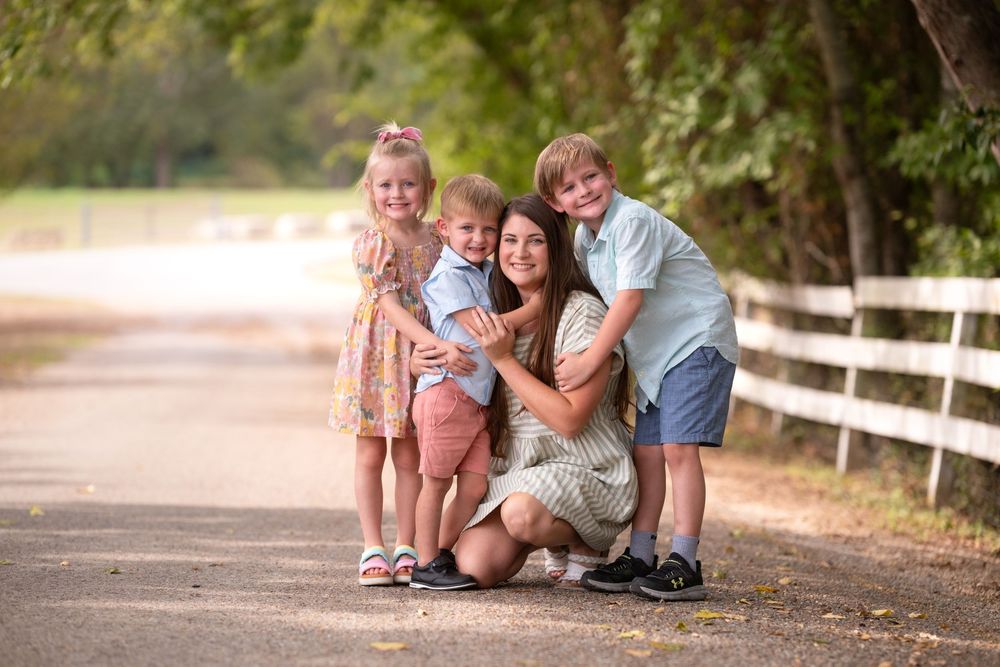 A smiling mother kneels on a tree-lined path with her three young children, posing for a warm family portrait outdoors.