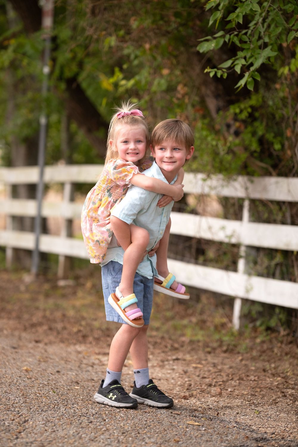 A young boy gives a smiling toddler girl a piggyback ride on a gravel path beside a white wooden fence.