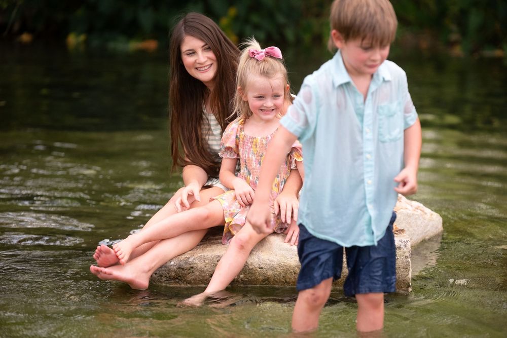 A woman and two young children sit and play on rocks in a shallow creek, smiling and enjoying the water outdoors.
