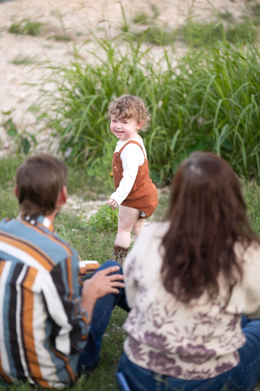 Happy toddler in brown overalls smiling at parents sitting on grass near tall green reeds outdoors.
