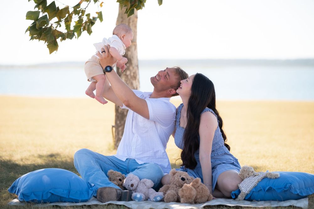 Family enjoying a sunny beach picnic with teddy bears, father lifting baby while mother smiles beside a tree.