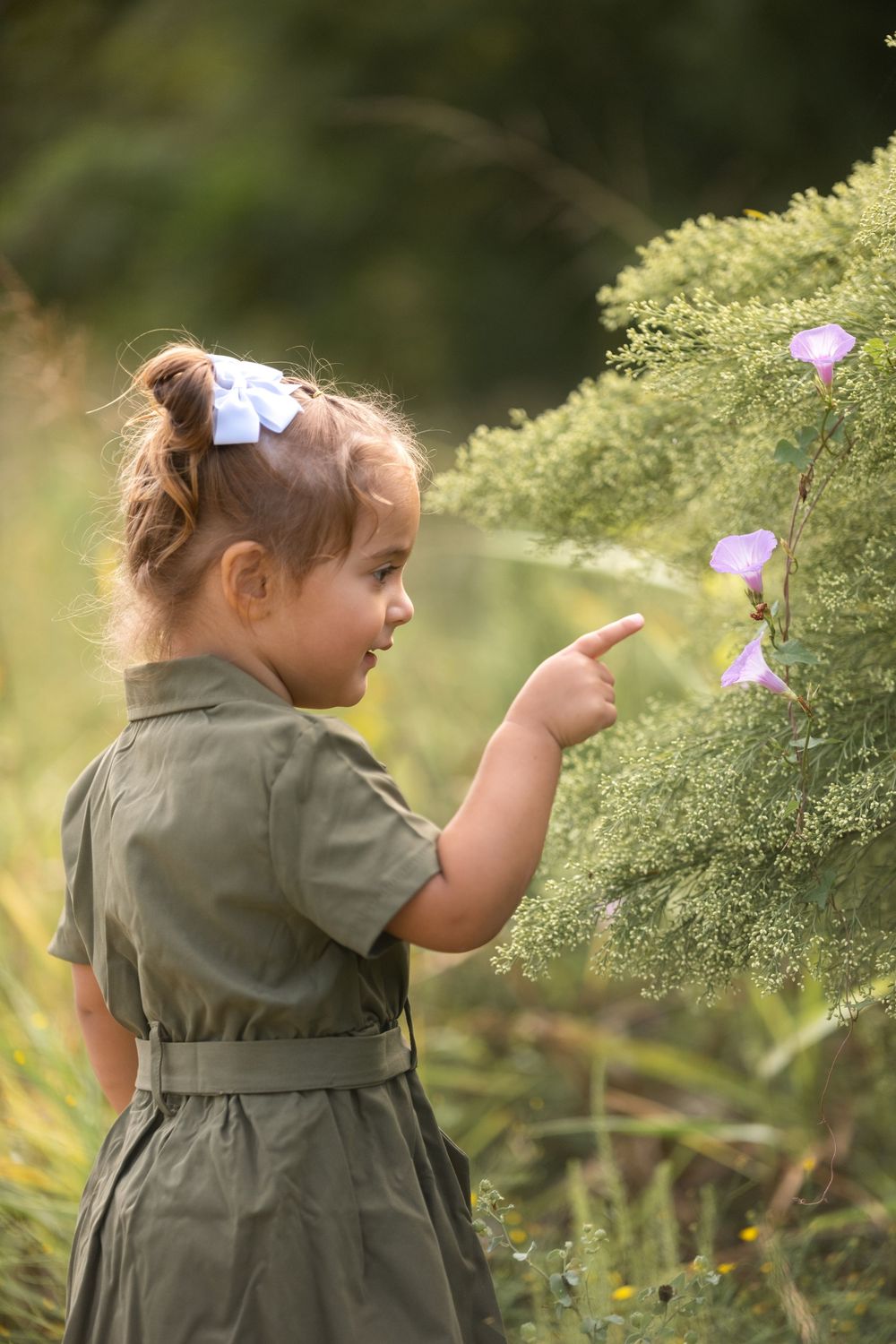 Young girl in olive dress pointing at pink wildflowers on a bush in a sunlit garden.