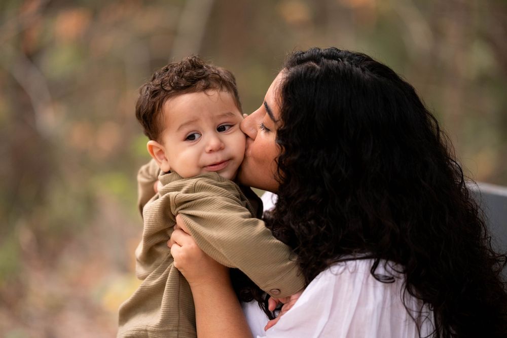 A mother tenderly kisses her smiling baby boy outdoors in a warm autumn setting.