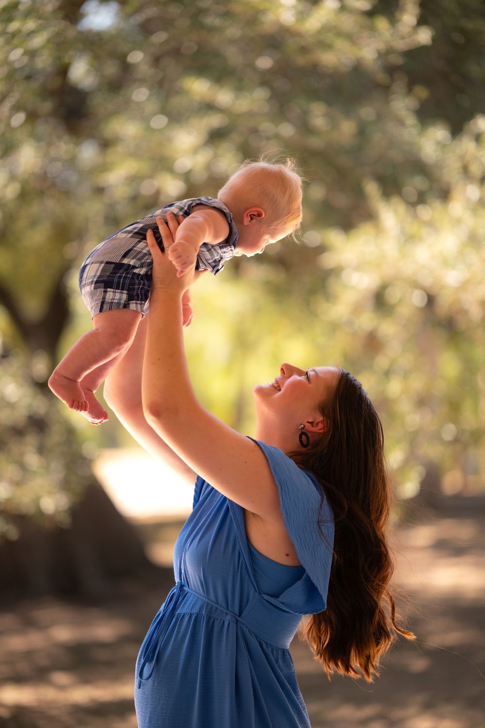 A mother in a blue dress joyfully lifts her baby overhead outdoors among green trees in warm sunlight.