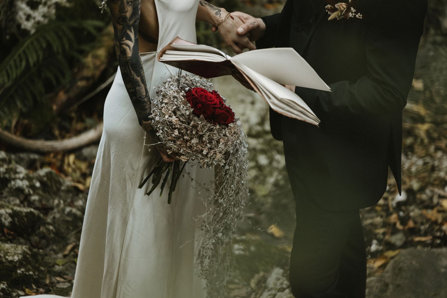 Bride holds a red rose bouquet with cascading greenery during outdoor ceremony.