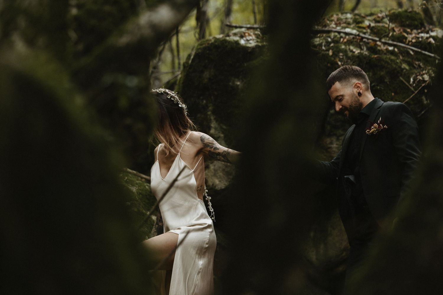 Bride and groom share a tender moment among mossy rocks in a dark forest.