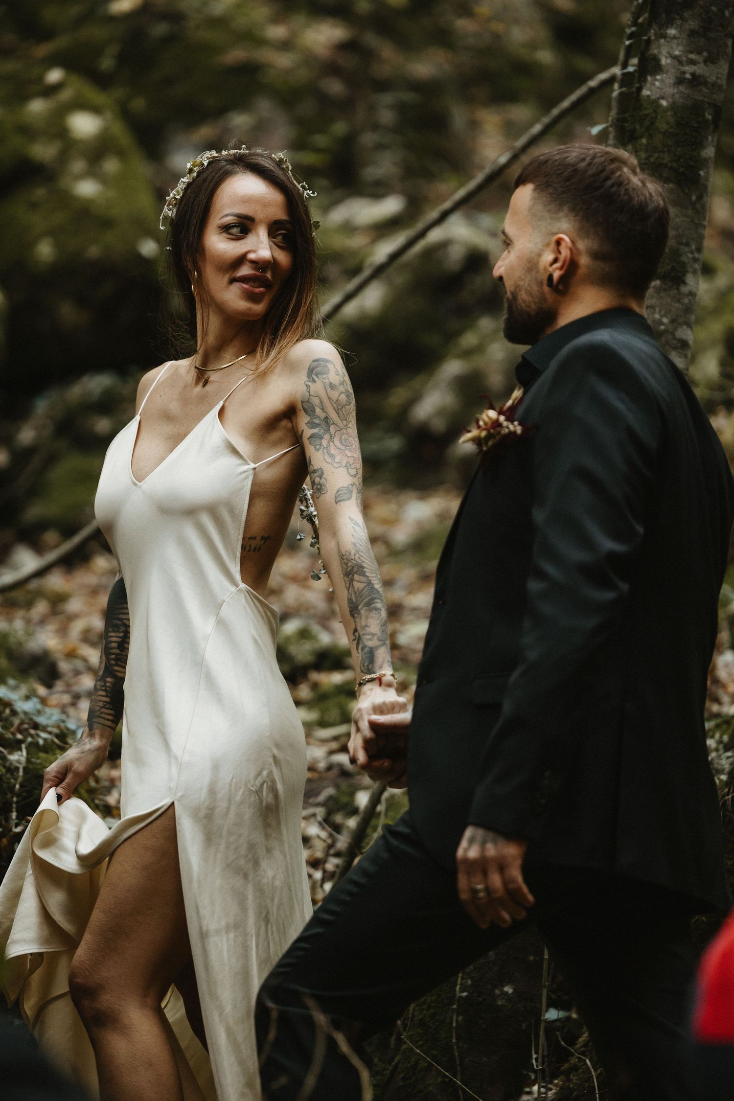 Tattooed bride in white slip dress with flower crown holds groom's hand walking through forest.