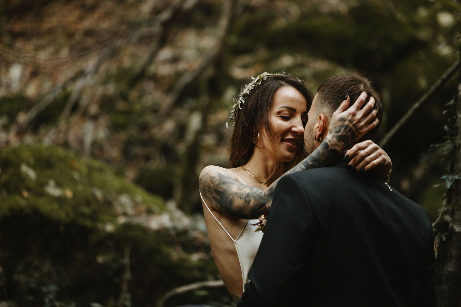 Tattooed bride and groom embrace intimately among mossy rocks in woodland wedding setting.