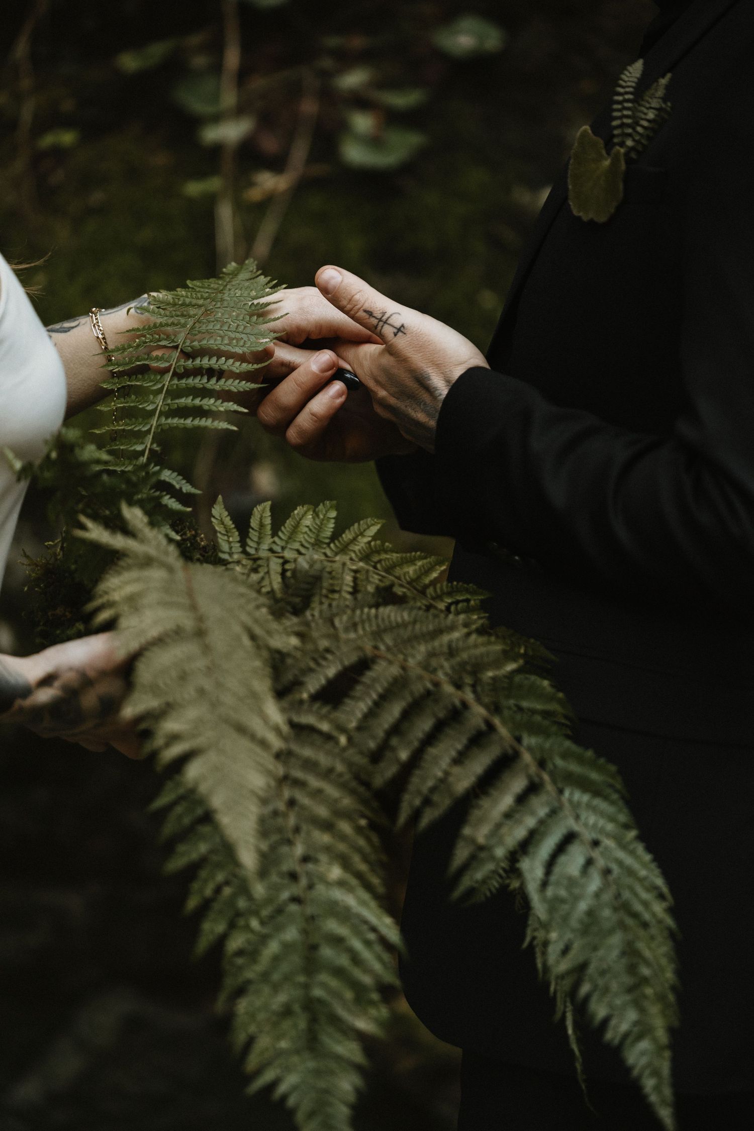 Bride and groom hold hands over lush green fern fronds during woodland wedding ceremony.
