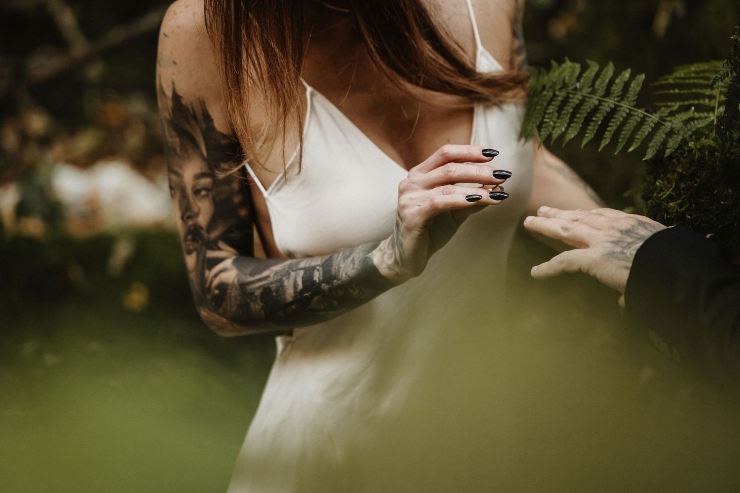 Close-up of tattooed bride's arm in white dress holding groom's hand amid green foliage.