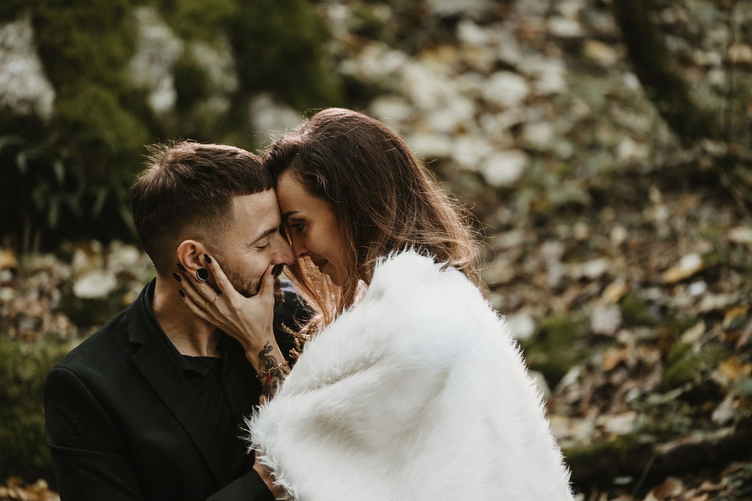 Laughing bride and groom share a tender moment wrapped in white fur in autumn woodland.