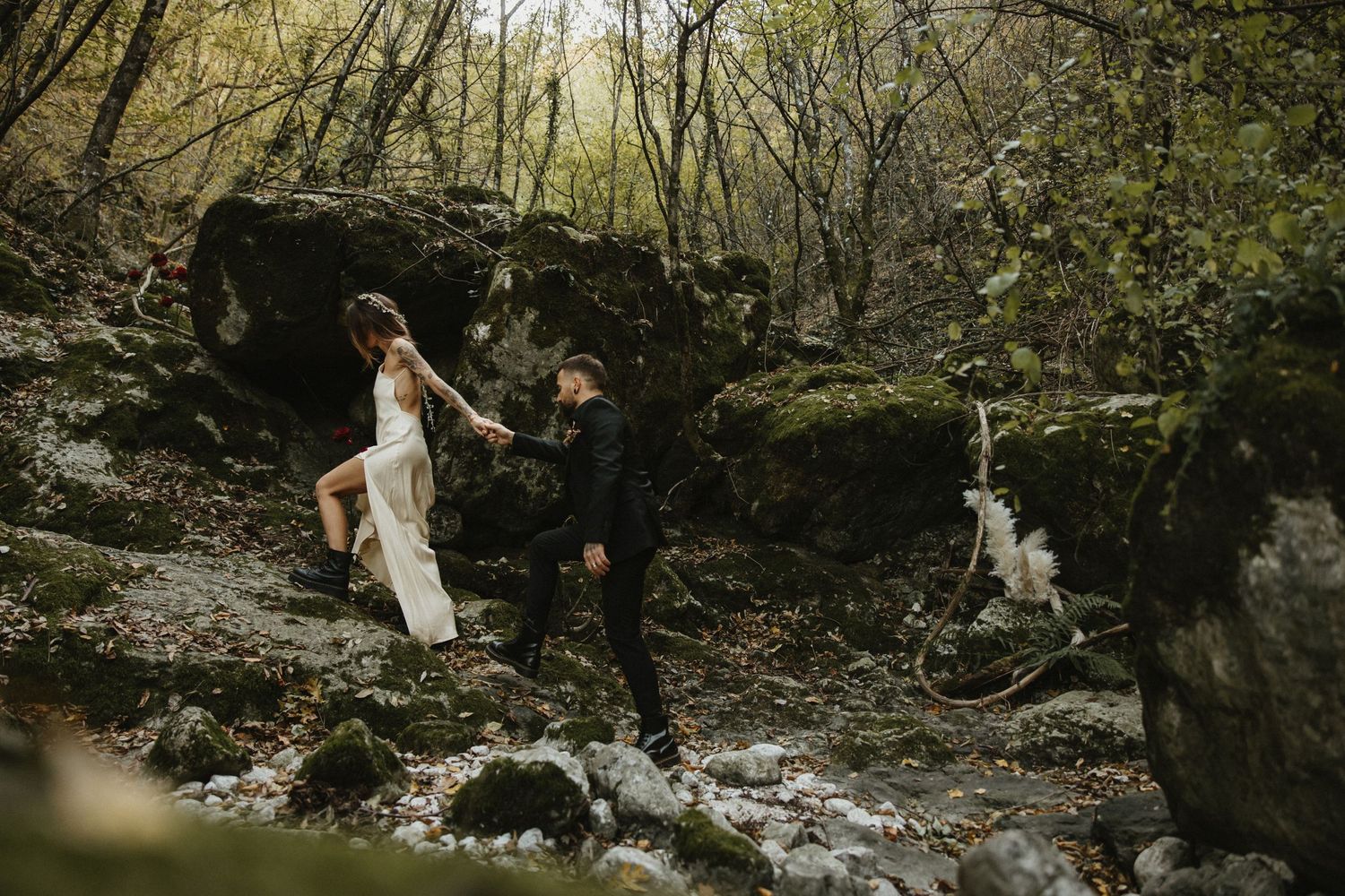 Bride and groom hold hands while crossing a rocky stream in a forest.