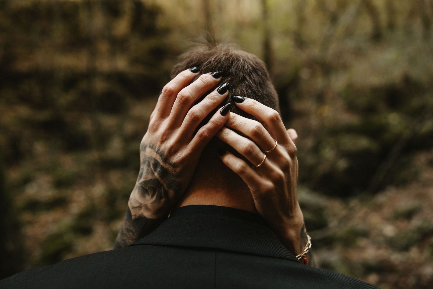 Tattooed hands with black nails holding the back of a mans head in a forest setting.